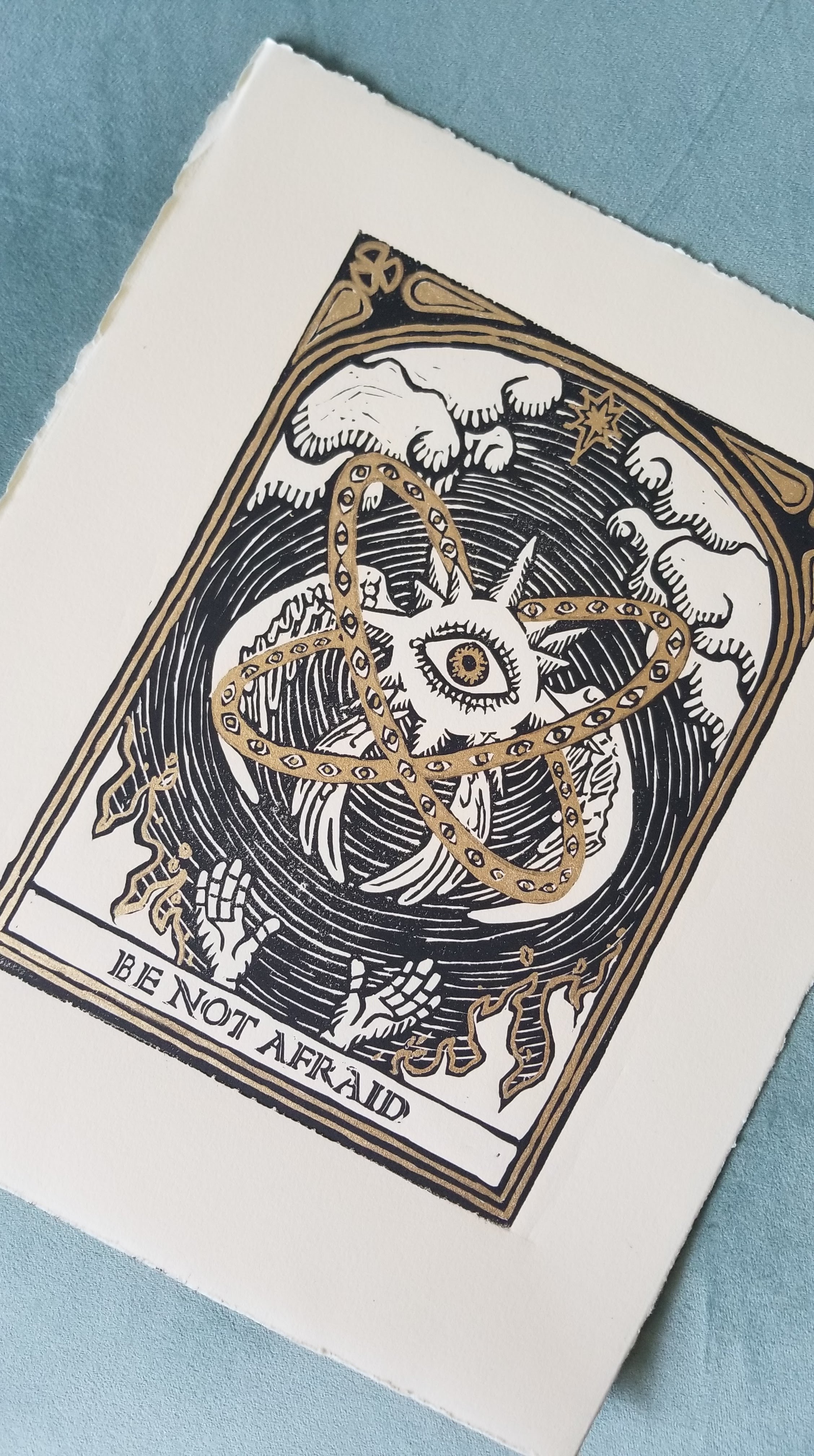 An angel flies above a pair of outstretched hands while fire burns. The print's title says "Be Not Afraid." The illustration includes gold foil accents.